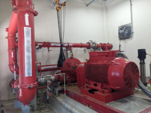 Sistema de bomba de combate a incêndio com tubulações vermelhas, motor elétrico e conjunto hidráulico instalado em sala técnica.